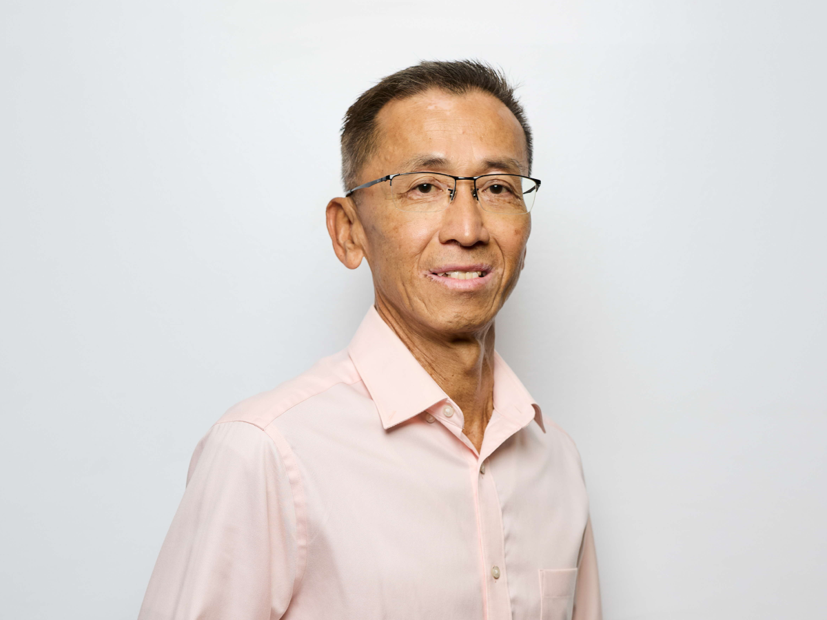 Mr Anwar Abdullah in a light pink shirt smiling and standing with hands together against a light grey background